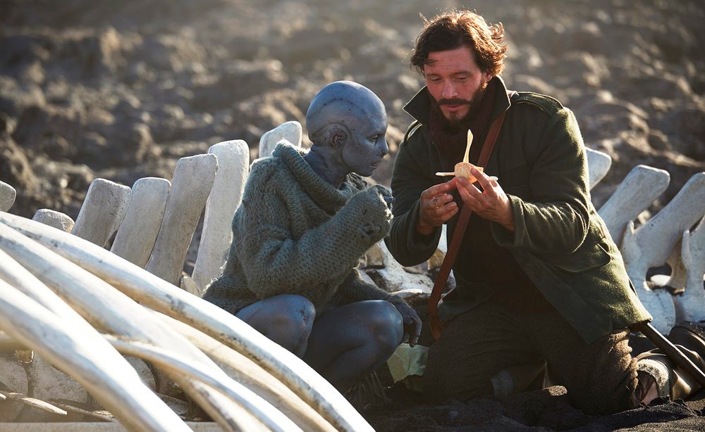 David as Friend in Cold Skin, kneeling among whale bones on a beach, showing a small carved figure to a grey-skinned creature