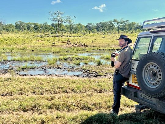 David leaning on a Painted Dog Conservation vehicle, watching a pack of painted dogs at a waterhole in Hwange National Park